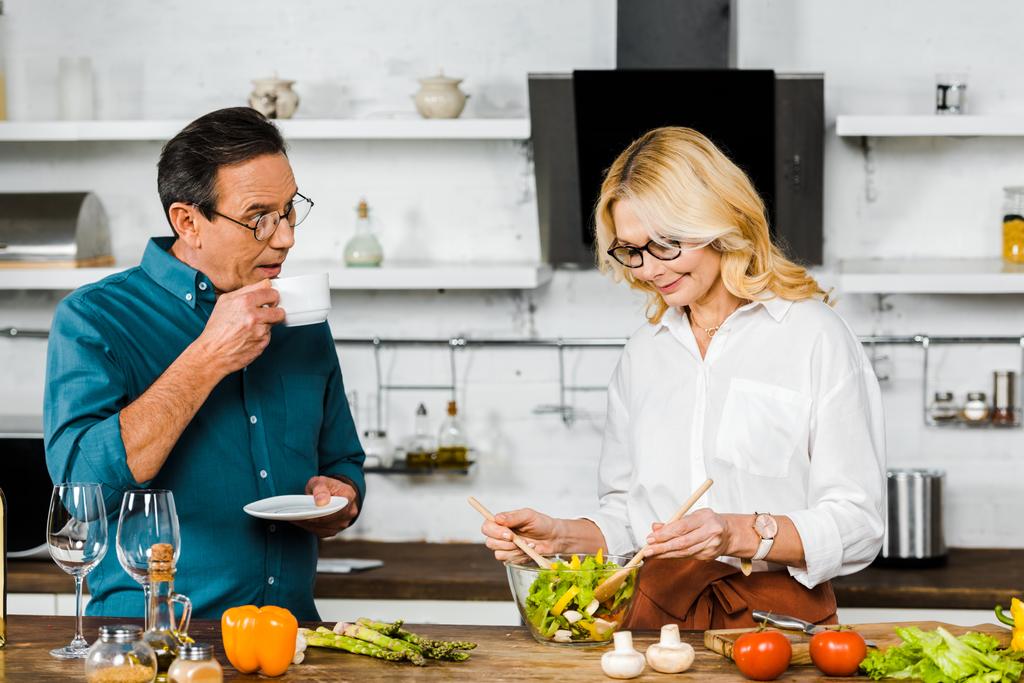 atraktivní starší žena míchání salát a manžel pít čaj v kuchyni - Fotografie, Obrázek