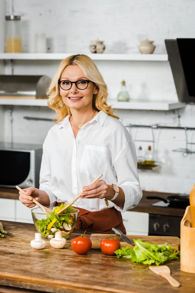 uśmiechający się piękne średniej wieku kobieta mieszania sałatki w szklanej misce w kuchni i patrząc na kamery - Zdjęcie, obraz