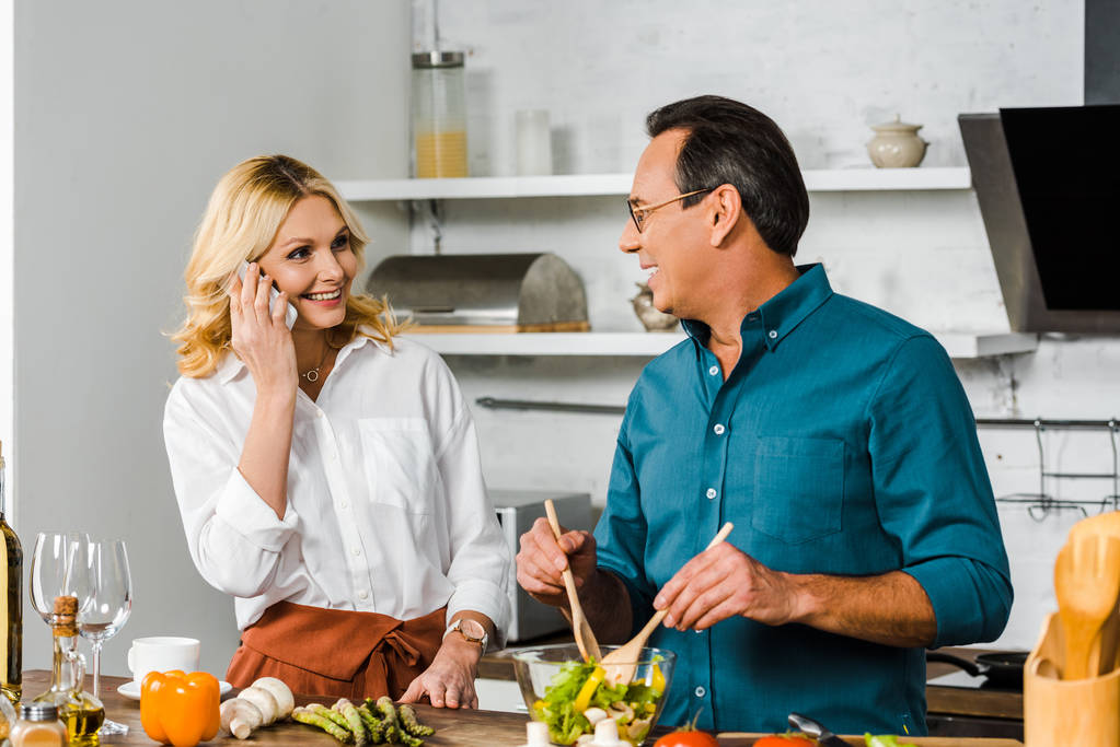 rijpe vrouw praten door smartphone en echtgenoot mengen salade in kom in keuken - Foto, afbeelding