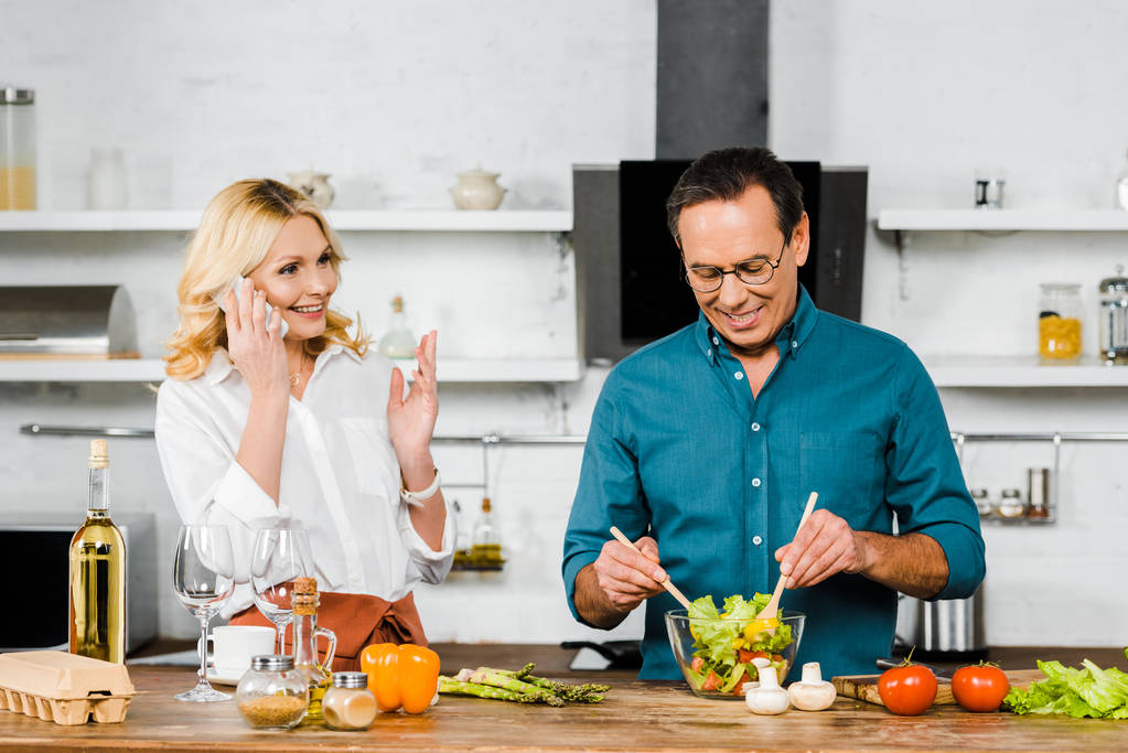 attrayant mature femme parler par smartphone et beau mari mélange salade dans un bol dans la cuisine
 - Photo, image