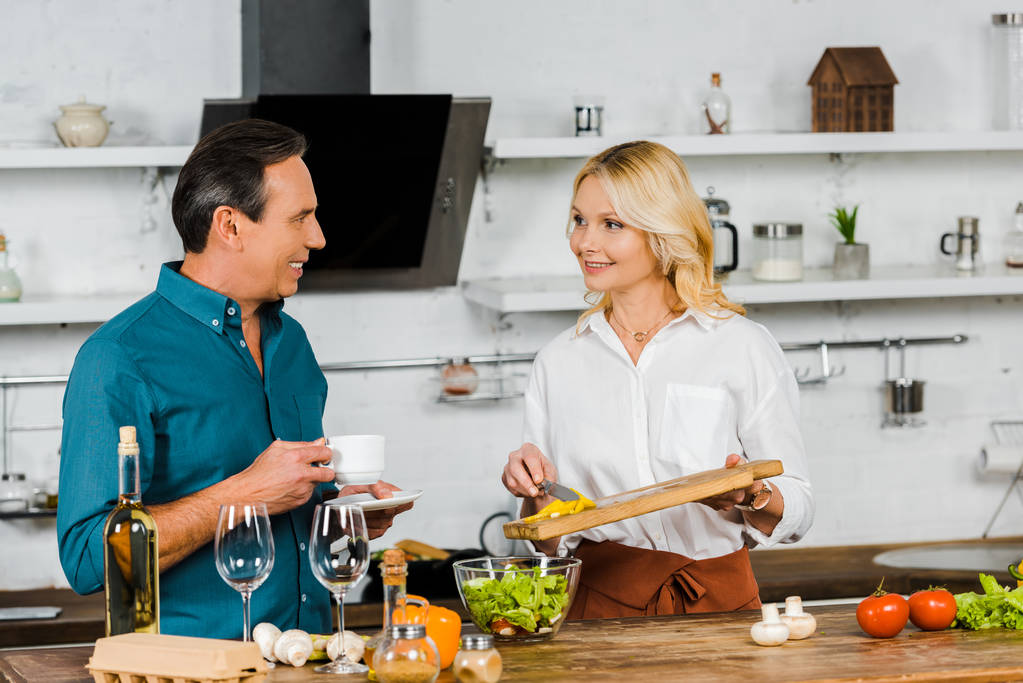 lächelnd attraktive reife Frau legt Gemüse in Schüssel und Ehemann hält Tasse Tee in der Küche - Foto, Bild