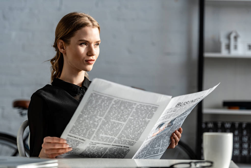 mooie zakenvrouw in zwarte formele slijtage zit aan Bureau en het lezen van de krant op werkplek - Foto, afbeelding