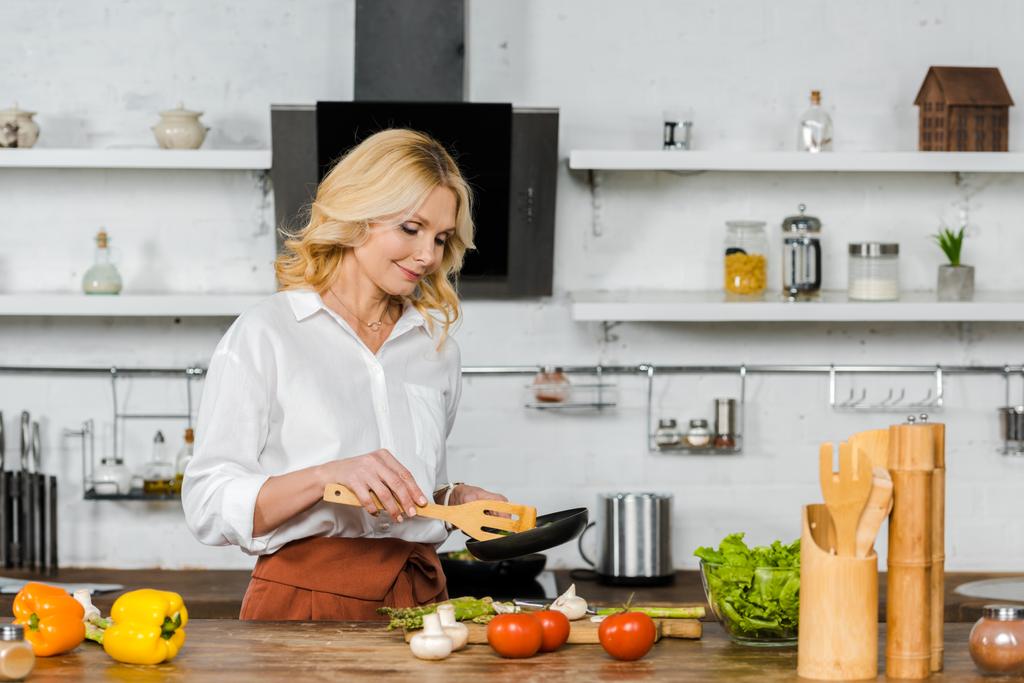 atraktivní středního věku žena vaření zeleniny na pánvi v kuchyni - Fotografie, Obrázek