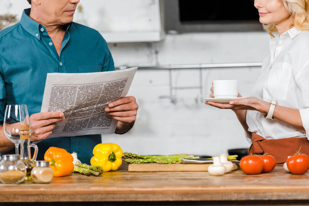 cropped image of mature wife holding cup of tea and husband reading newspaper while cooking in kitchen - Photo, Image