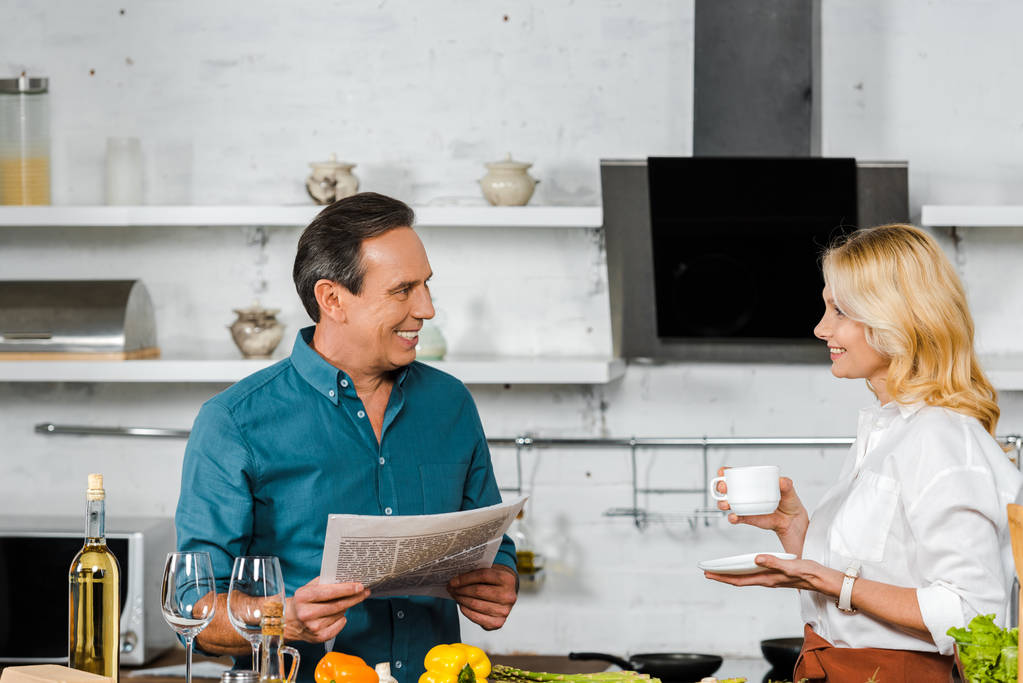 紅茶、キッチンで料理をしながら新聞を読んで夫の成熟した妻ホールディング カップを笑顔 - 写真・画像