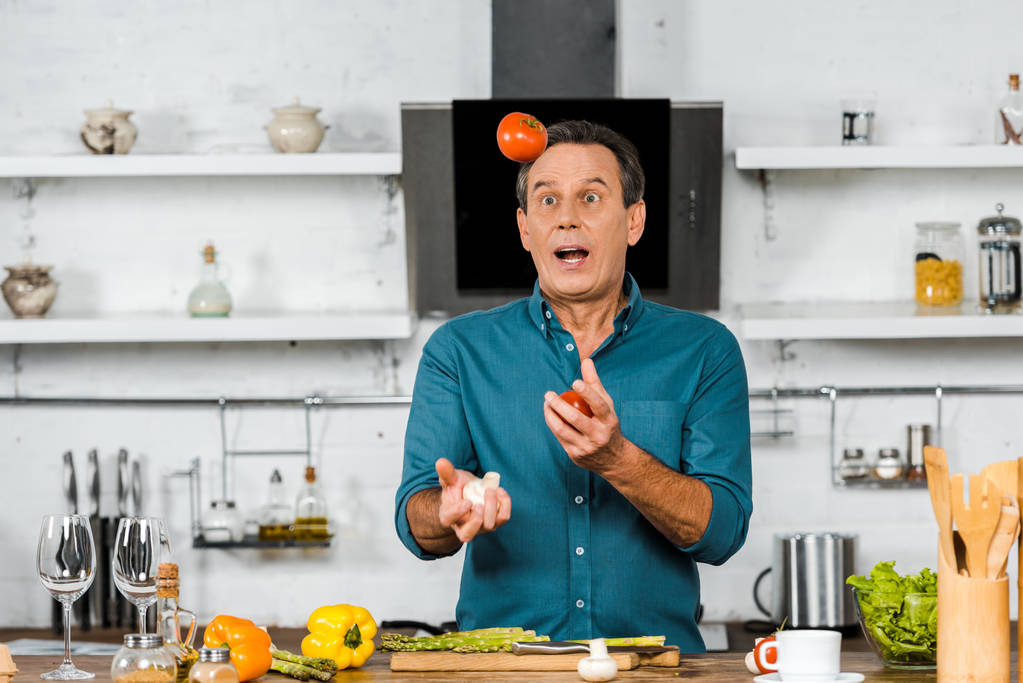 schöner Mann mittleren Alters beim Kochen in der Küche mit Gemüse jongliert überrascht - Foto, Bild