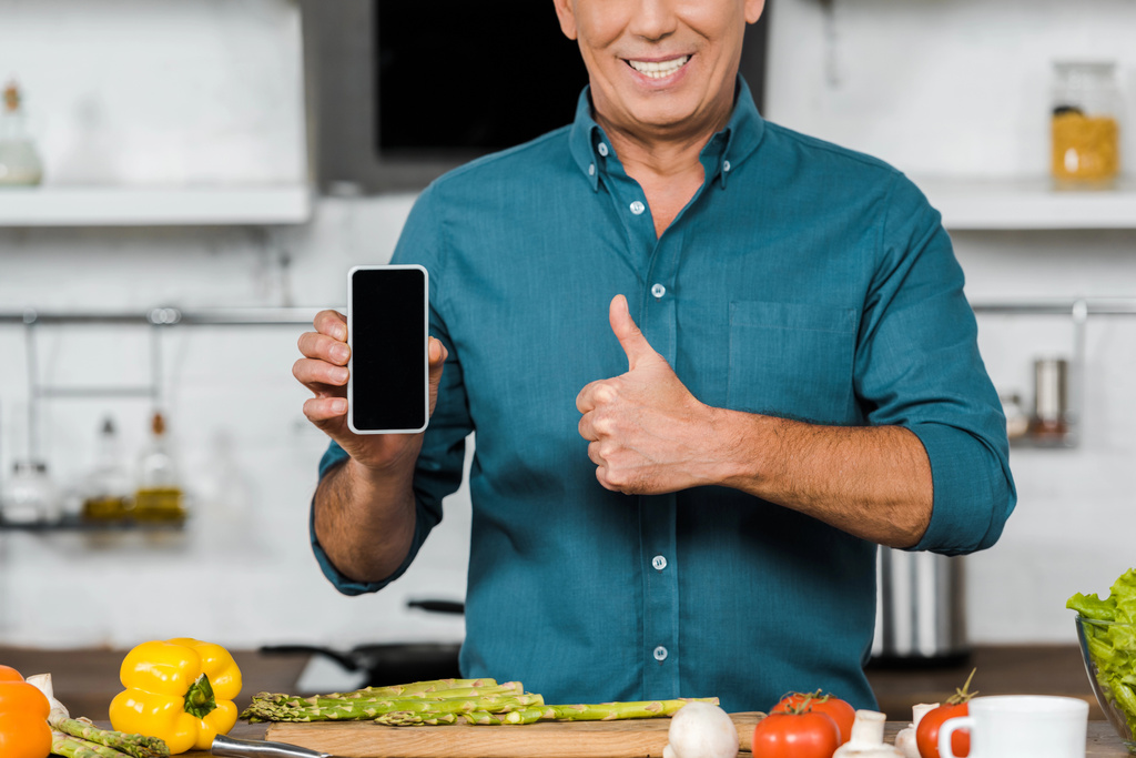 περικομμένη εικόνα χαμογελώντας όμορφος μέσης ηλικίας άτομο που κρατά το smartphone με την κενή οθόνη και δείχνει τον αντίχειρα επάνω στην κουζίνα - Φωτογραφία, εικόνα