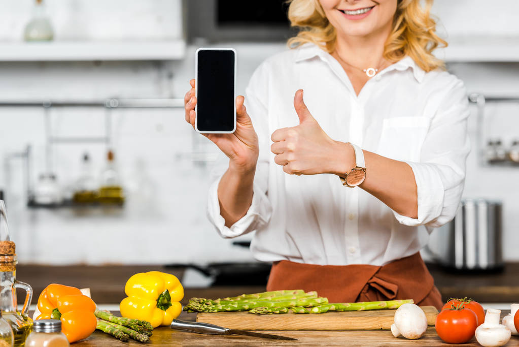 oříznutý obraz středního věku žena držící smartphone s prázdnou obrazovkou a ukazuje palcem v kuchyni - Fotografie, Obrázek