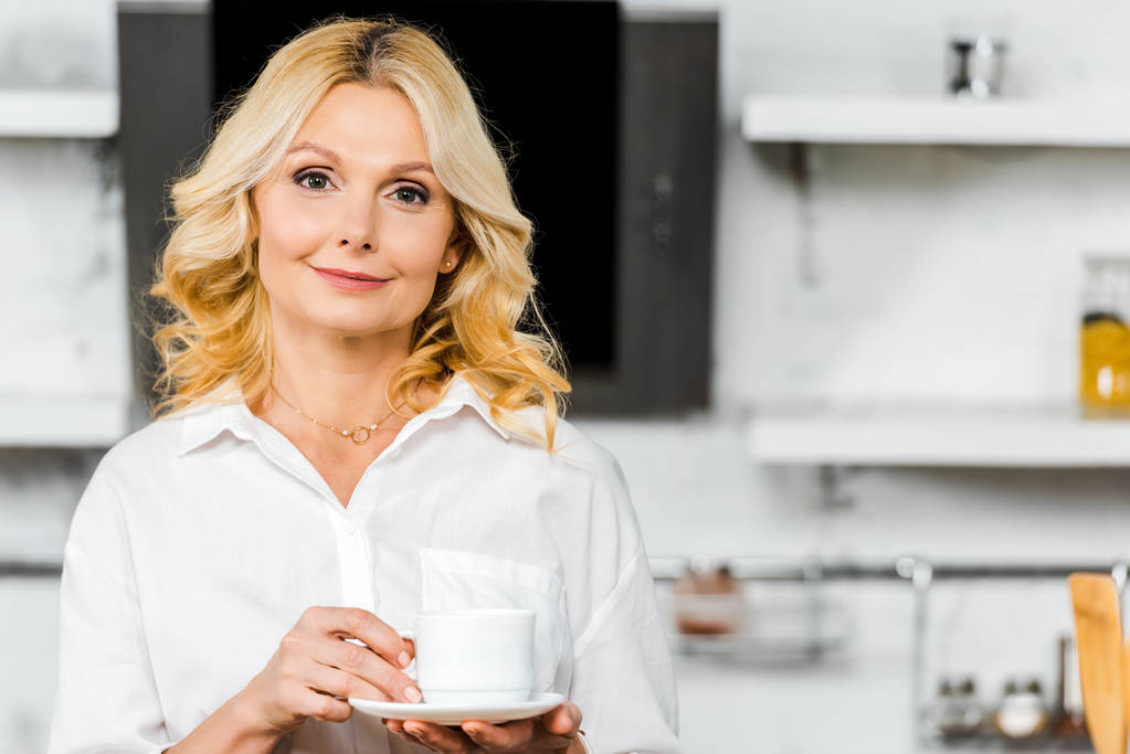 attractive middle aged woman holding cup of tea in kitchen and looking at camera - Photo, Image