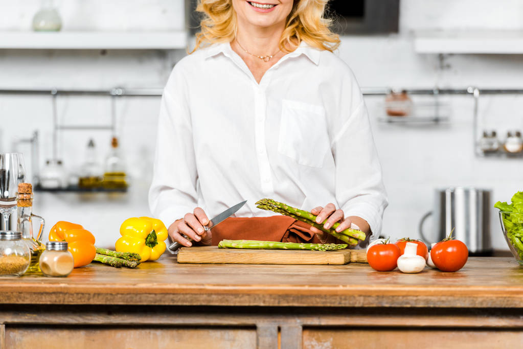 rajattu kuva hymyilevä keski-ikäinen nainen leikkaa vihanneksia keittiössä
 - Valokuva, kuva