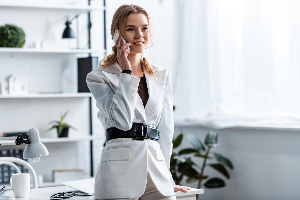 Lächelnde Geschäftsfrau in weißer Festkleidung spricht am Arbeitsplatz auf dem Smartphone - Foto, Bild