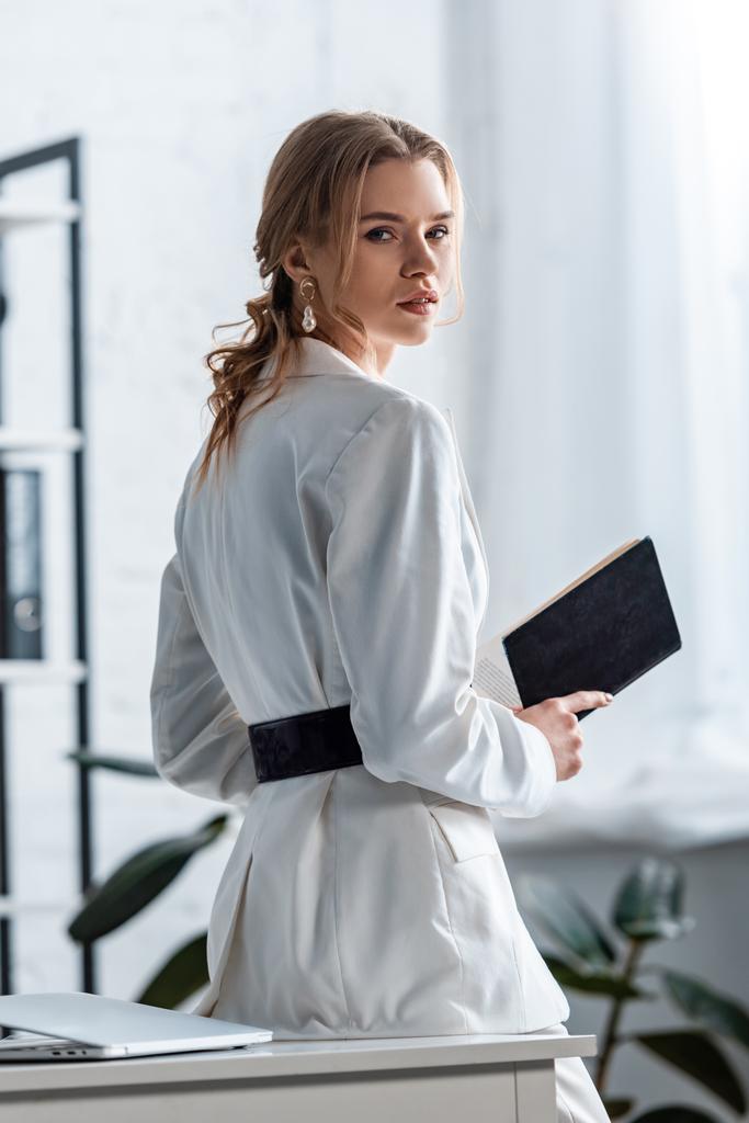 krásná podnikatelka v oblasti formální oblečení drží notebook a při pohledu kamery na pracovišti - Fotografie, Obrázek