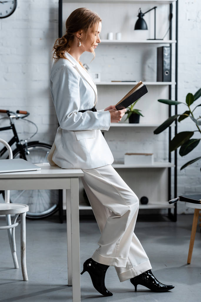femme d'affaires élégante en tenue formelle assis sur le bureau et le cahier de lecture sur le lieu de travail
 - Photo, image