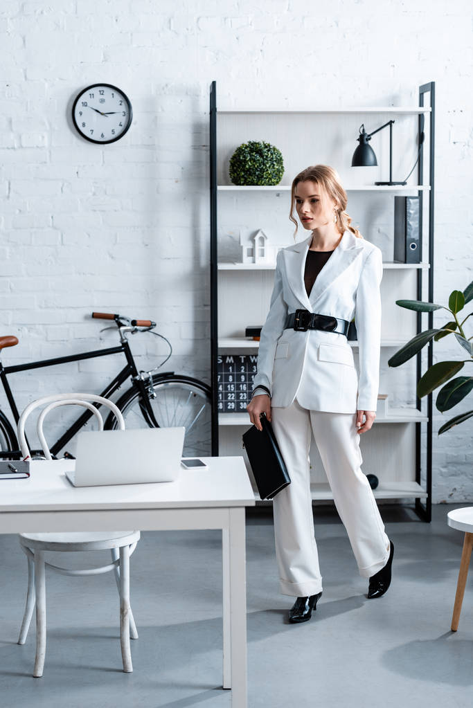 серйозна бізнес-леді в білому формальному носінні тримає блокнот в сучасному офісному інтер'єрі
 - Фото, зображення