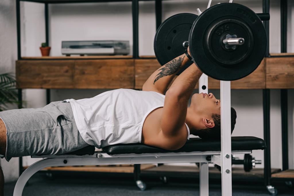 tattooed mixed race man exercising with barbell in home gym  - Photo, Image