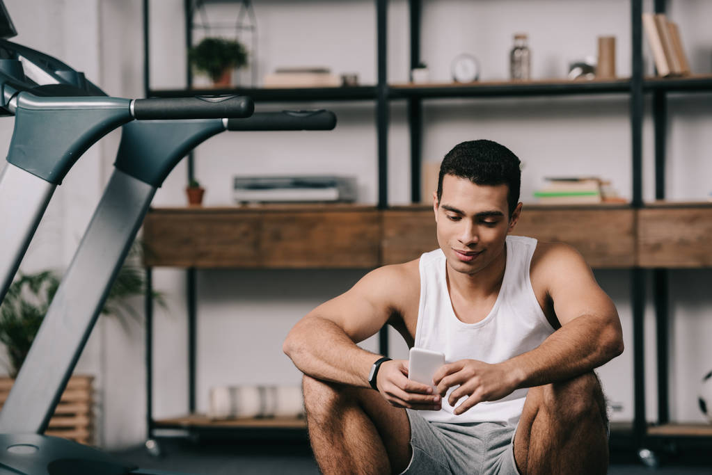 schöner zweirassiger Sportler, der neben dem Laufband sitzt und das Smartphone benutzt - Foto, Bild