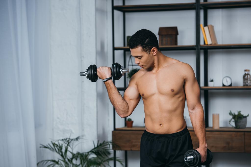 schöner Mann mit zwei Rassen, der beim Training mit Kurzhanteln im Fitnessstudio auf die Muskeln schaut  - Foto, Bild