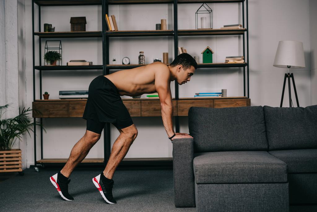 musculoso hombre de raza mixta haciendo ejercicio cerca de sofá en sala de estar
  - Foto, Imagen