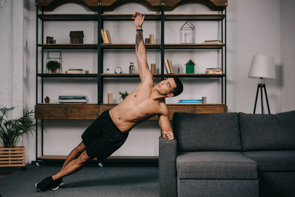 knappe getatoeëerd gemengd ras man doen plank oefening in woonkamer  - Foto, afbeelding