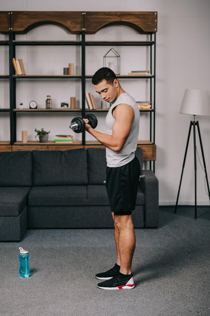 apuesto hombre de raza mixta mirando los músculos mientras hace ejercicio en la sala de estar
 - Foto, Imagen