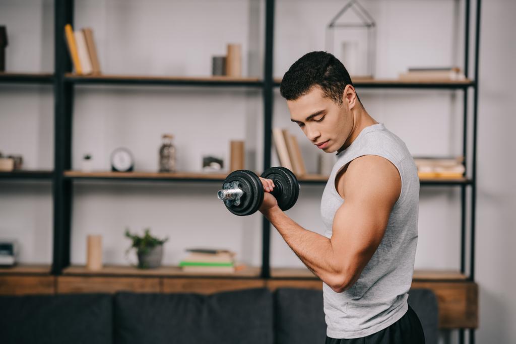 guapo hombre de raza mixta mirando los músculos en la mano mientras hace ejercicio con la mancuerna en la sala de estar
 - Foto, Imagen
