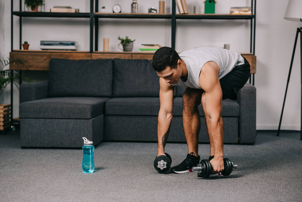 mixed race sportsman taking dumbbells from floor in living room - Photo, Image