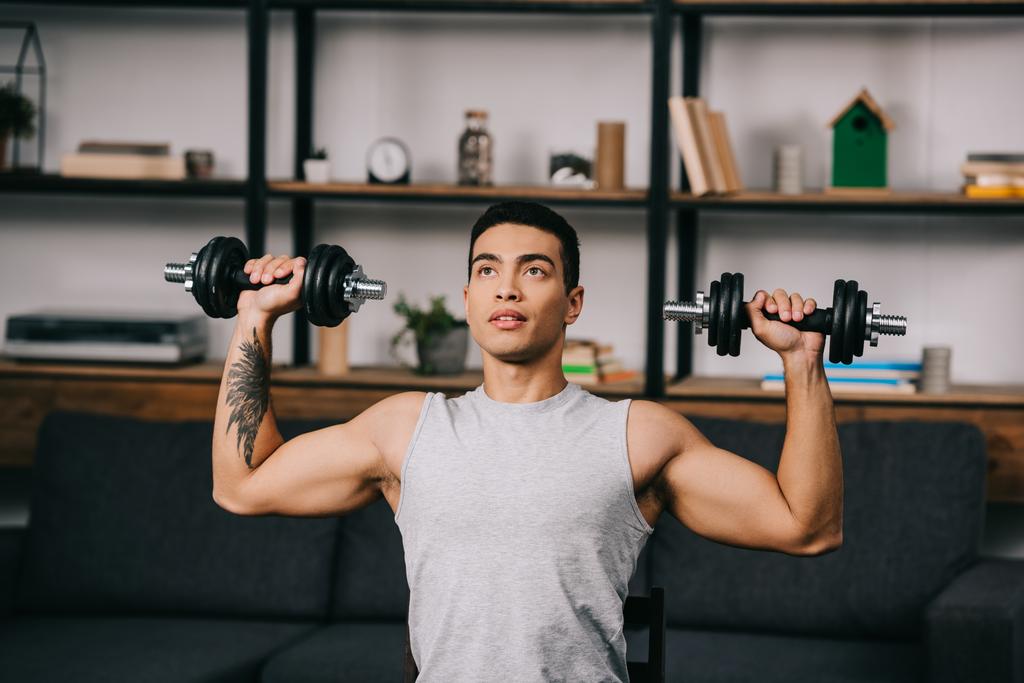 handsome mixed race athlete exercising with dumbbells in living room - Photo, Image