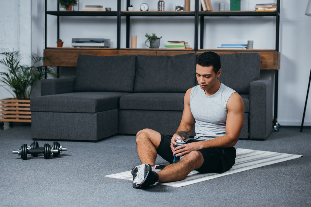 Mann sitzt mit Sportflasche auf Fitnessmatte im Wohnzimmer - Foto, Bild