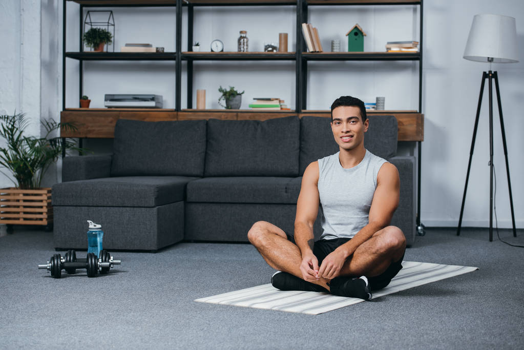 fröhlicher Mann mit zwei Rassen sitzt auf Fitnessmatte im Wohnzimmer  - Foto, Bild