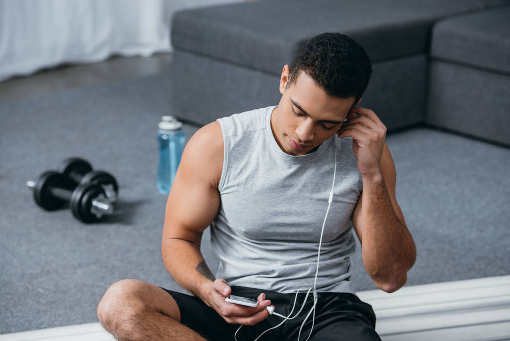 Selektywny fokus bi rasowe człowieka za pomocą smartfona i słuchanie muzyki, siedząc na fitness Mata - Zdjęcie, obraz
