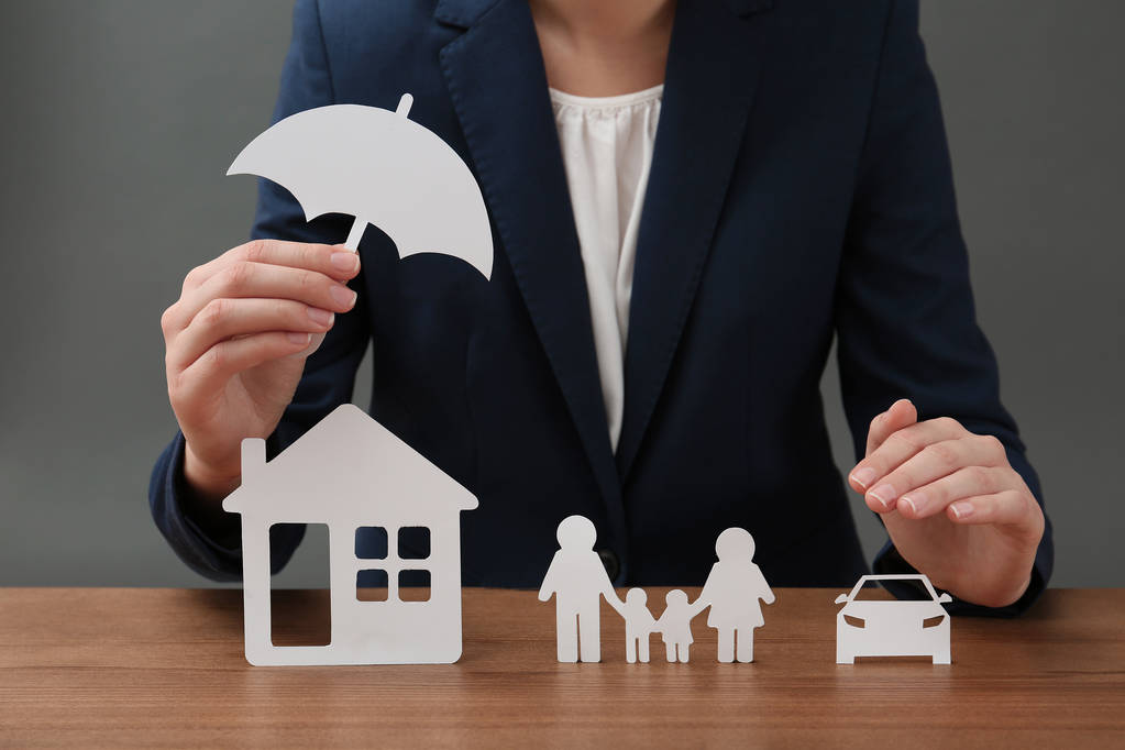 Mujer con familia de papel recortado, casa, coche y paraguas en la mesa, primer plano. Concepto seguro de vida
 - Foto, Imagen