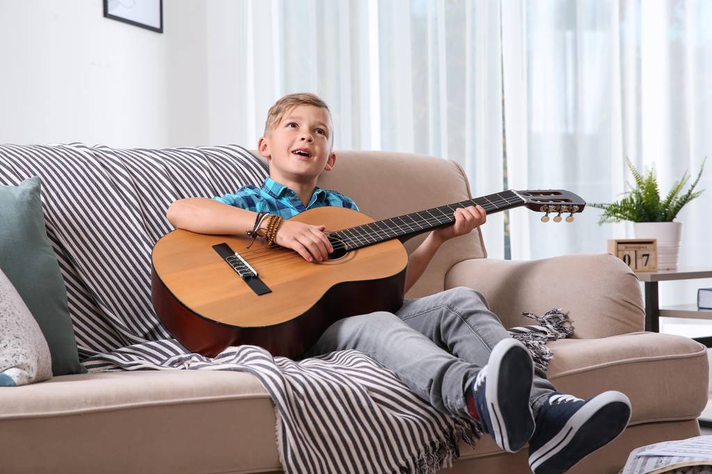 Ładny mały chłopiec gra na gitarze na kanapie w pokoju - Zdjęcie, obraz