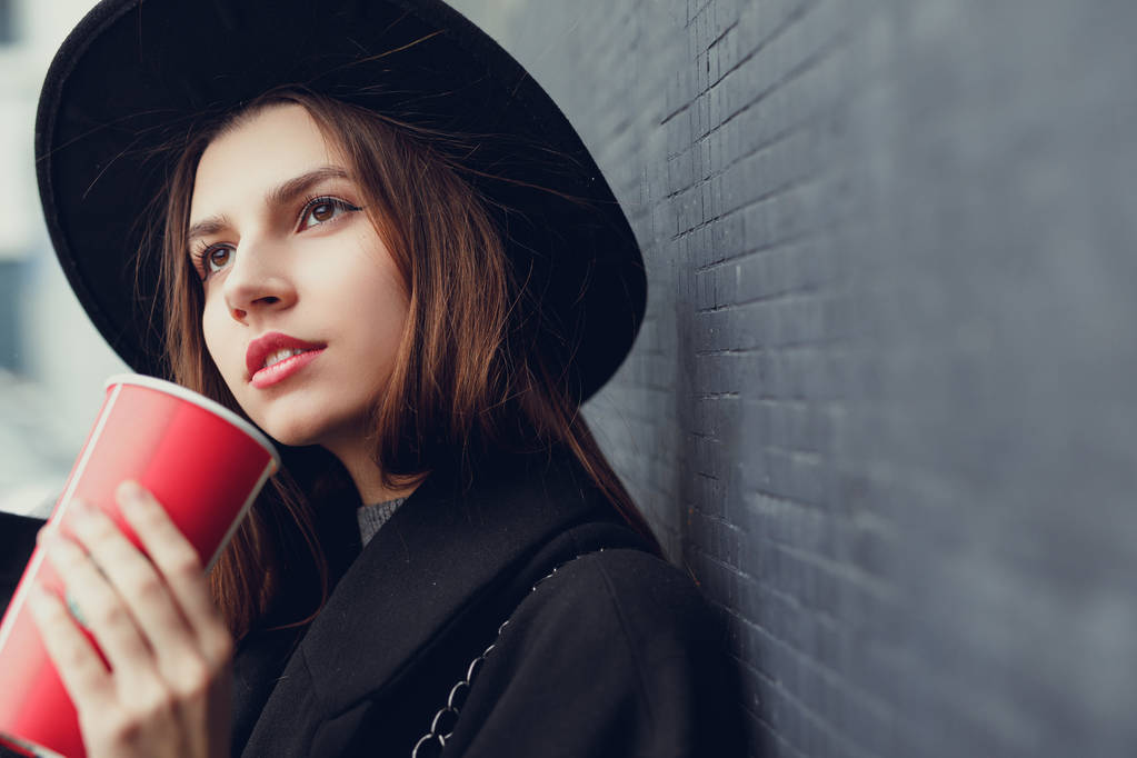Joven mujer de la moda sostener su taza caliente de café, de pie cerca de la pared llevar sombrero negro mirar a su alrededor y sonreír a la cámara. Copiar espacio. - Imagen
 - Foto, Imagen