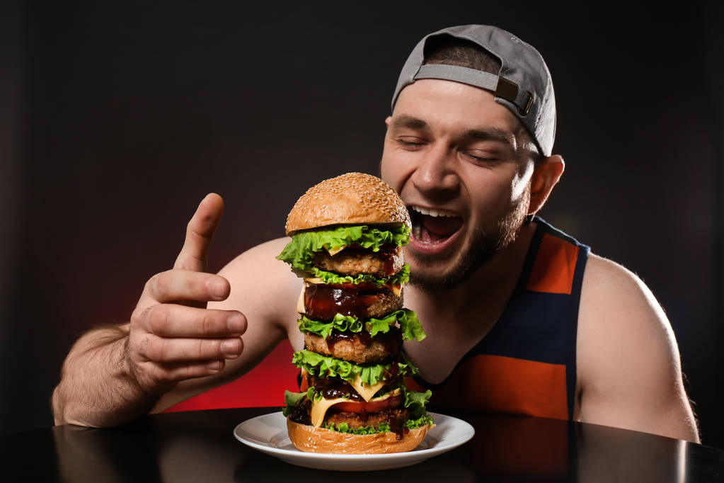 Молодий голодний чоловік їсть величезний бургер на чорному тлі
 - Фото, зображення
