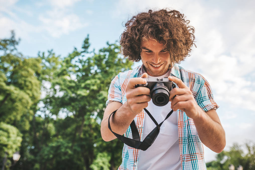 Κάτω shot δείτε Καυκάσιος άνδρα με σγουρά μαλλιά ελέγχοντας φωτογραφίες με την ψηφιακή φωτογραφική μηχανή. Νεαρό όμορφος άνδρας φοράει πουκάμισο, με ψηφιακή φωτογραφία κάμερα στέκεται στο φόντο της φύσης. Άνθρωποι, ταξίδια - Φωτογραφία, εικόνα