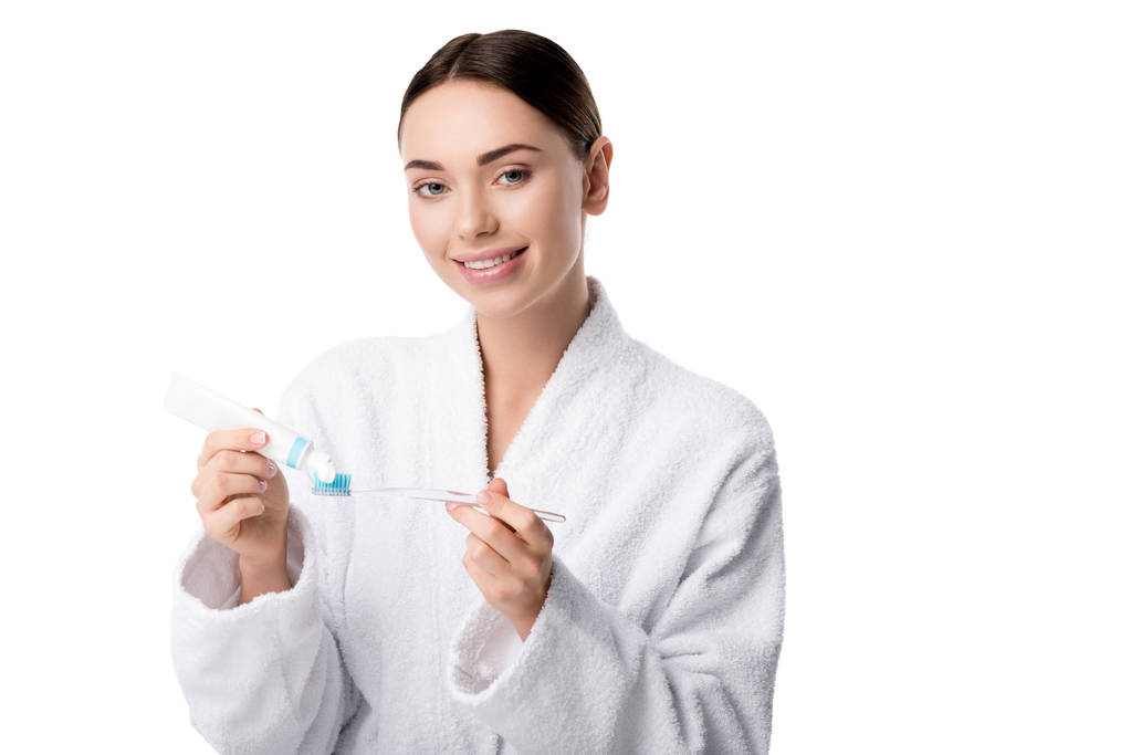 mujer sonriente en albornoz blanco poniendo pasta de dientes en el cepillo de dientes aislado en blanco
 - Foto, Imagen
