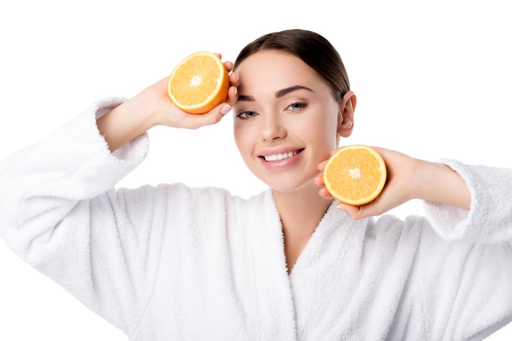 mooie lachende vrouw in witte badjas holding sinaasappelen en kijken naar camera geïsoleerd op wit - Foto, afbeelding