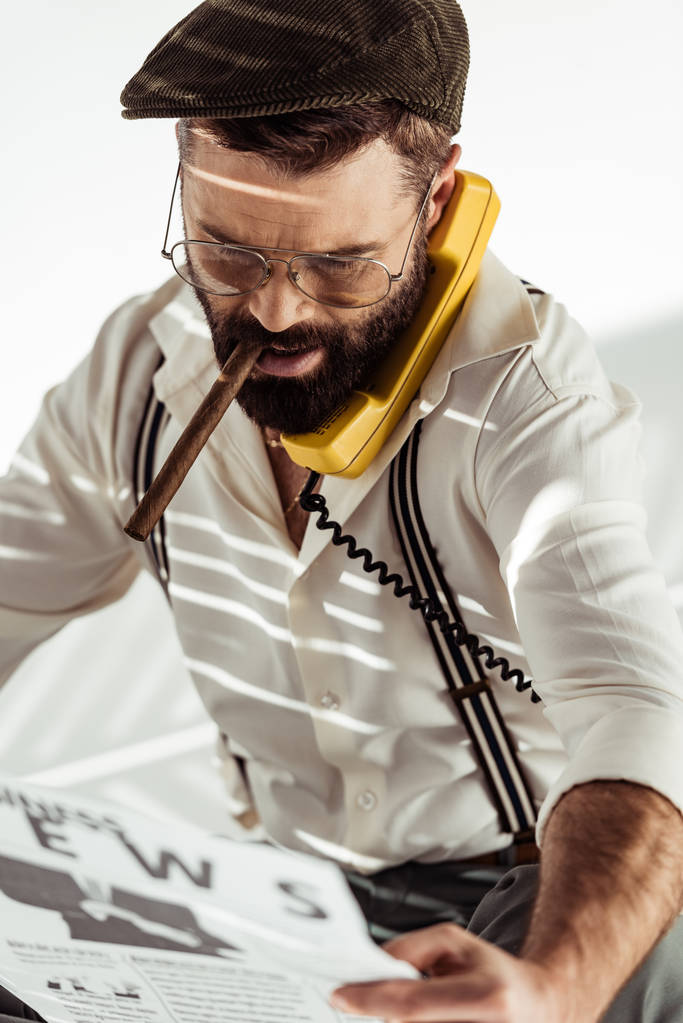 vousatý muž v brýlích a čepici mluví o vinobraní telefonu, kouřil doutník a čtenářský deník - Fotografie, Obrázek