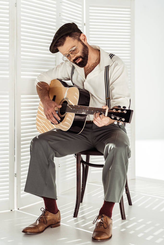 hezký vousatý hudebník v čepici sedí na židli a hrál na akustickou kytaru  - Fotografie, Obrázek