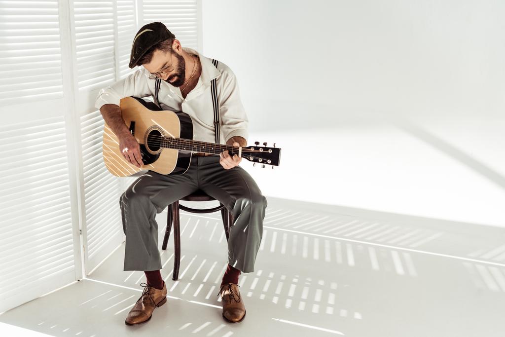 όμορφος γενειοφόρος άνδρας στο καπάκι κάθεται σε καρέκλα και να παίζει κιθάρα κοντά στο λευκό δωμάτιο διαχωριστικό - Φωτογραφία, εικόνα