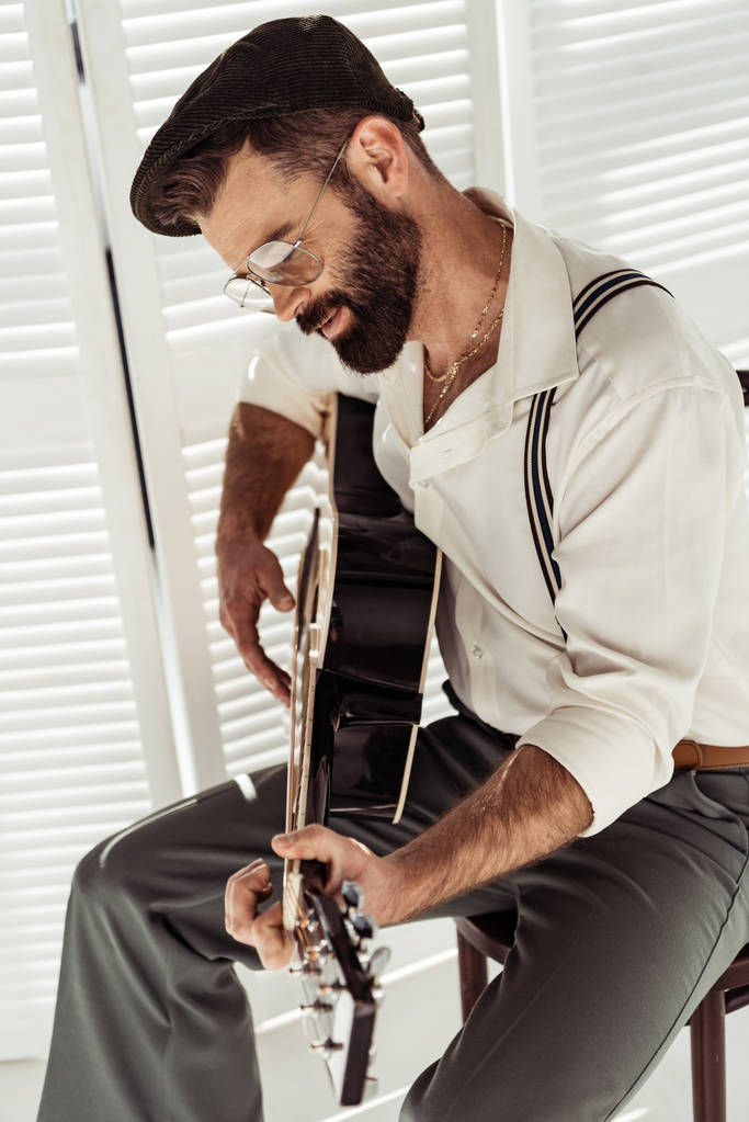 красивий бородатий чоловік сидить на стільці і грає на акустичній гітарі біля розкладного екрану в кімнаті
 - Фото, зображення