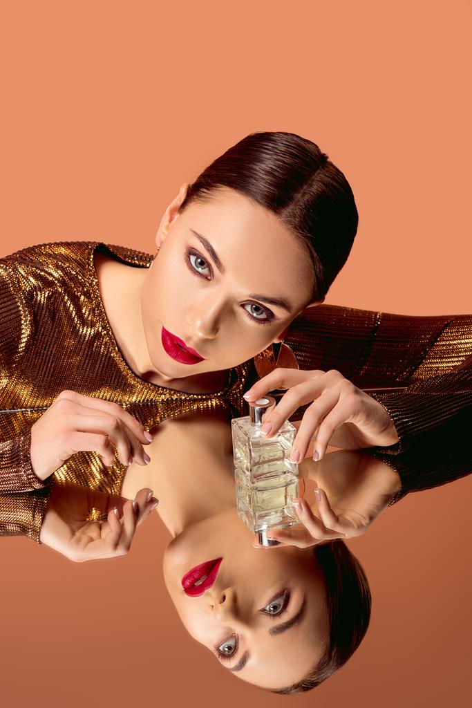 mujer atractiva en ropa dorada con maquillaje glamoroso, botella de perfume y espejo reflexión posando aislado en naranja
 - Foto, imagen