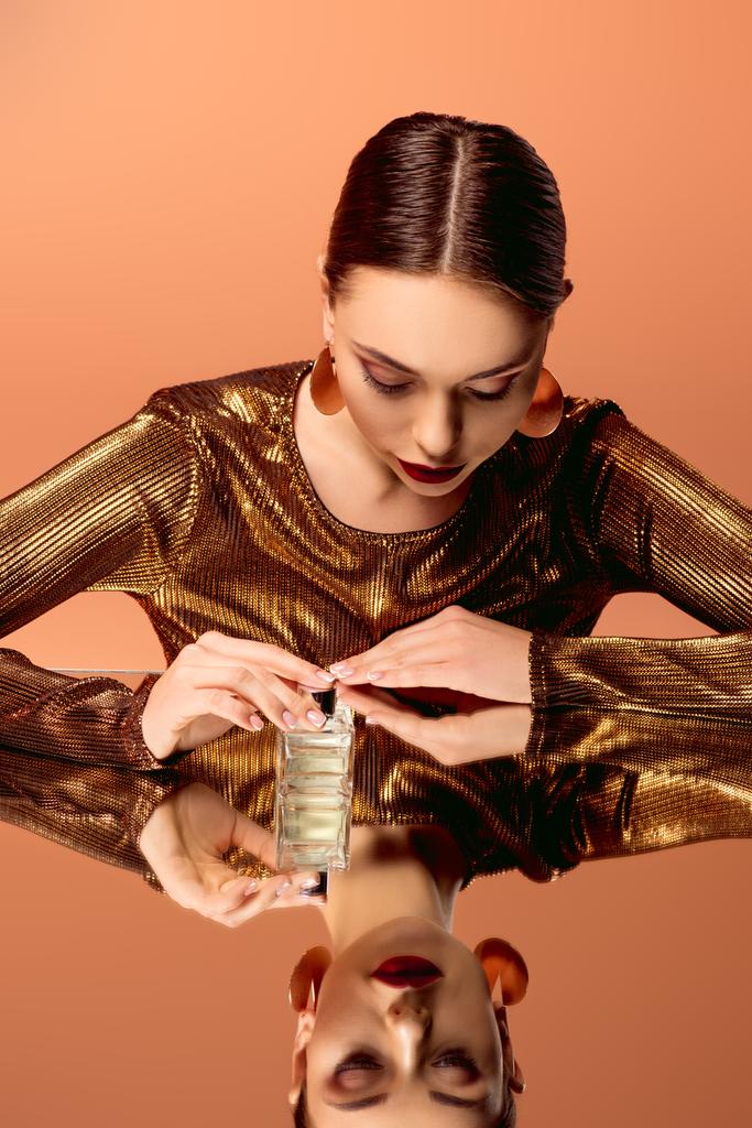 mooie vrouw in gouden kleren met parfum fles en spiegel reflectie geïsoleerd op oranje - Foto, afbeelding