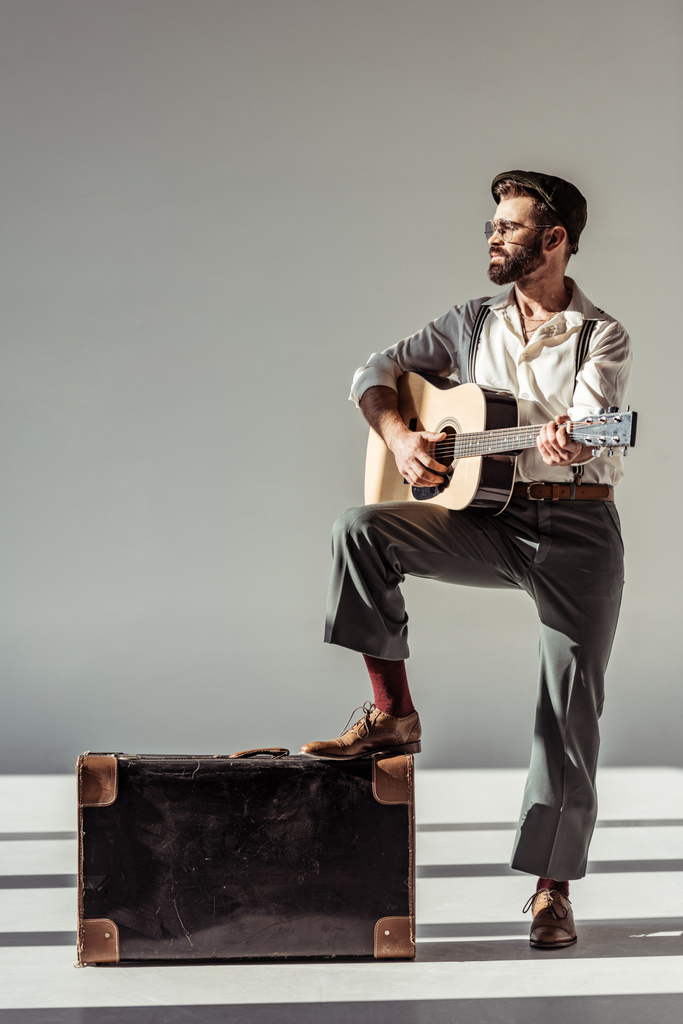 músico barbudo em boné perto de mala vintage tocando guitarra acústica em fundo cinza
 - Foto, Imagem