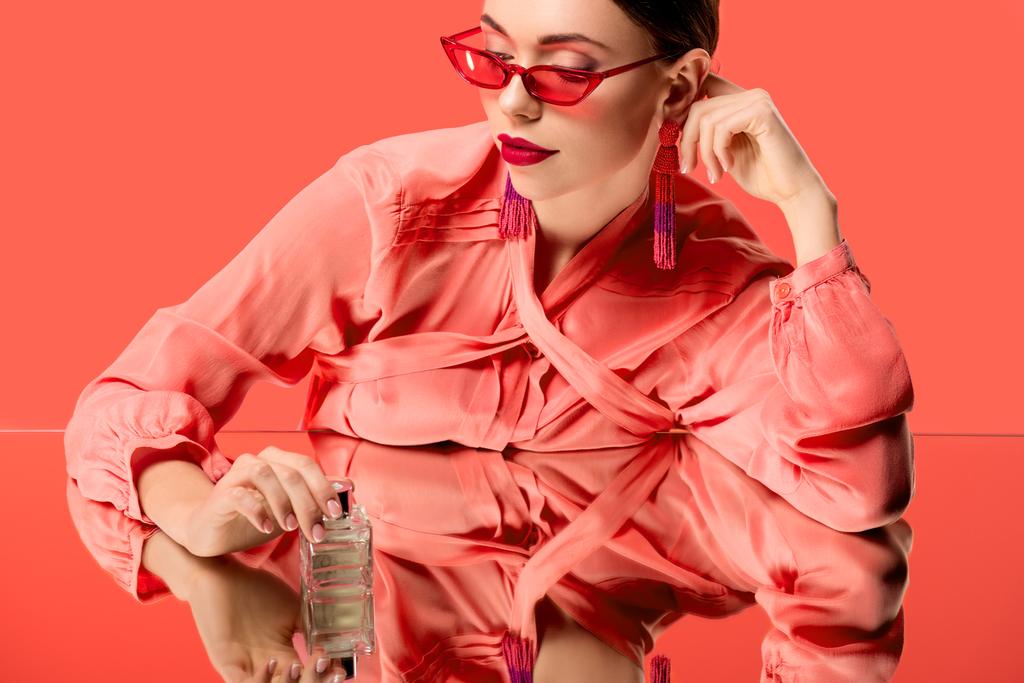 κομψή γυναίκα σε μπλούζα και κόκκινα γυαλιά ηλίου που ποζάρει με Άρωμα μπουκάλι και καθρέπτης αντανάκλασης απομονώνονται σε κοραλλιογενείς ζωής - Φωτογραφία, εικόνα