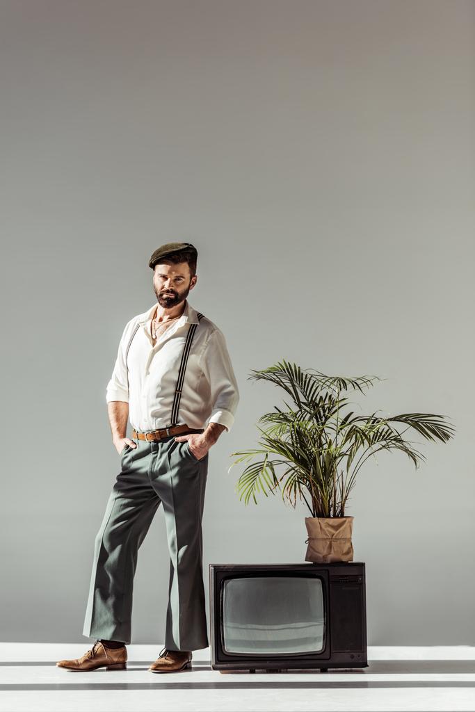 handsome bearded man in cap standing near vintage tv with plant in pot and looking at camera  - Photo, Image