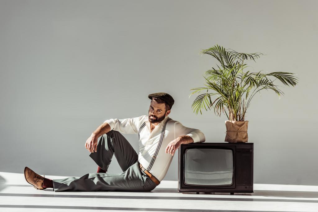 bonito barbudo homem no cap sentado no chão perto de vintage tv com planta no pote e olhando para a câmera
 - Foto, Imagem