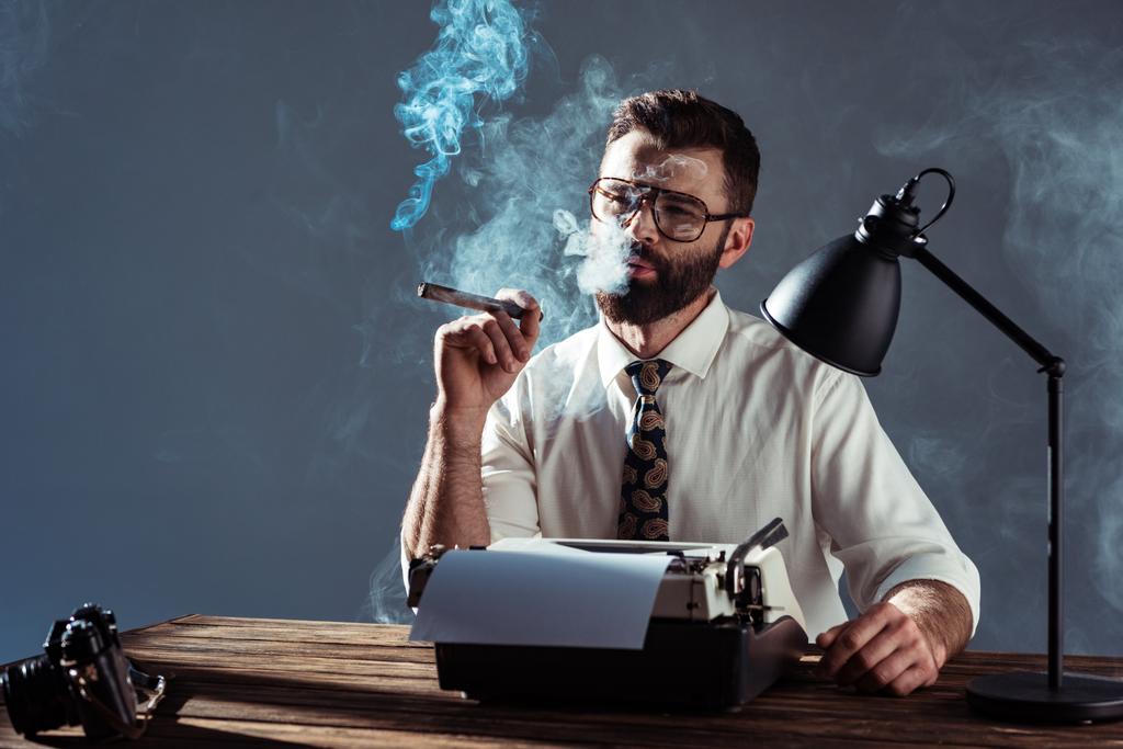 pensativo periodista barbudo sentado a la mesa con máquina de escribir y fumando sobre fondo gris
 - Foto, Imagen