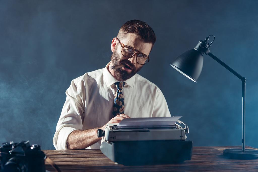 bearded journalist sitting at table, typing on typewriter, smoking and looking at camera on grey background - Photo, Image