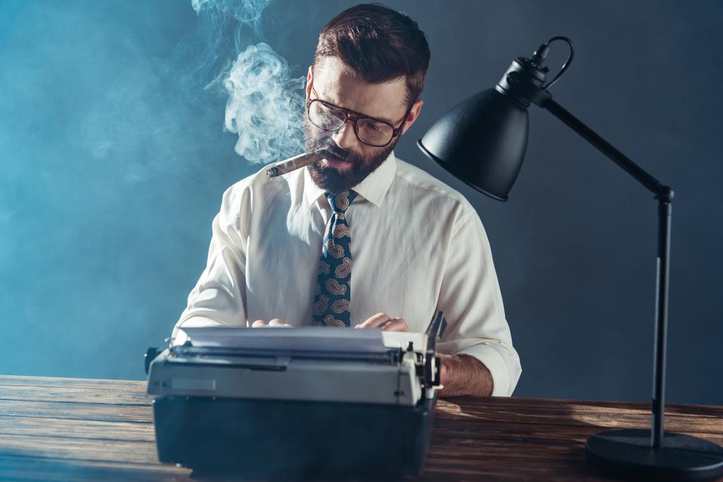 pohledný novinář v brýlích sedí u stolu, psaní na stroji vinobraní a kouření na šedém pozadí - Fotografie, Obrázek